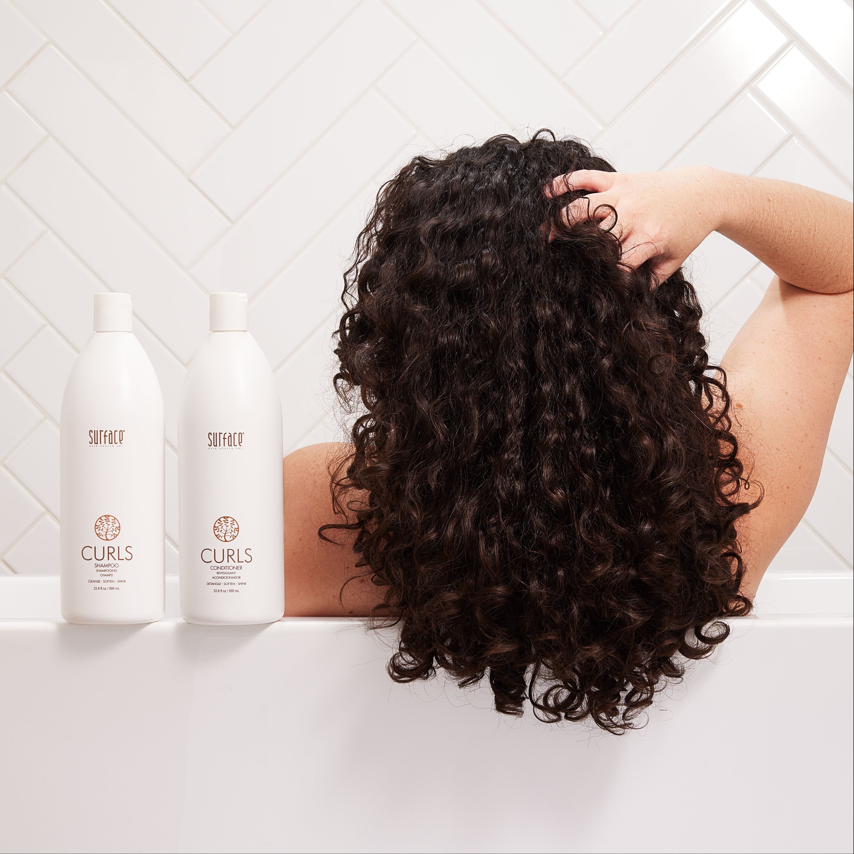 Person with curly hair in a bathtub with Curls Shampoo and Conditioner bottles on a white tiled wall background
